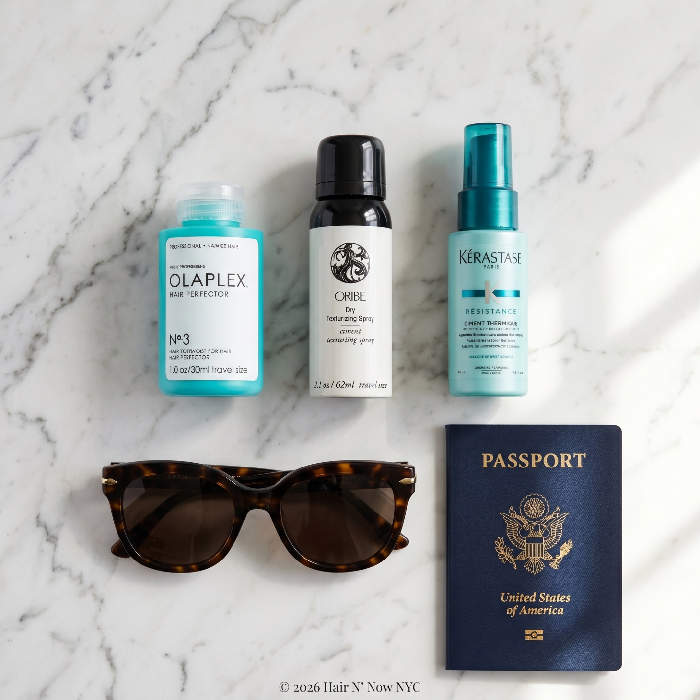 Flat lay of travel-size Oribe Dry Texturizing Spray, Olaplex No. 3, Kerastase Ciment Thermique, sunglasses, and passport.